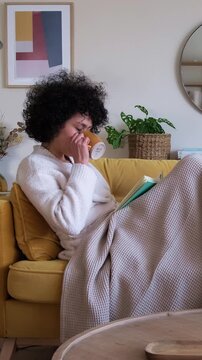 Relaxed woman with afro hair drinking tea and reading a book on the sofa