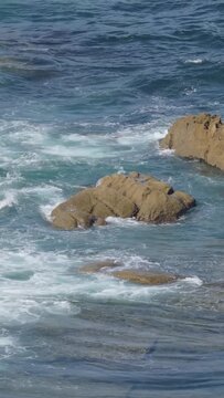 Dynamic vertical 9:16 video showing ocean waves crashing against rugged rocks along the coastline of Baiona, Spain.