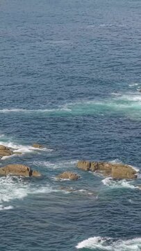 Vertical 9:16 video showing waves crashing against rocks along the coastline in Baiona, Spain. No people are present in the scene.