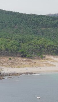 Vertical 9:16 aerial video of Praia de Melide beach in Cangas, Galicia, Spain. Scattered beachgoers and umbrellas are visible on the sand, while the lush green forest rises behind the beach.