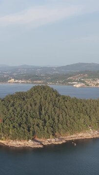 Vertical 9:16 aerial video showing a lush, forested island (Illa de Tambo) surrounded by calm blue waters, with the coastline and hills of Combarro, Spain visible in the distance.