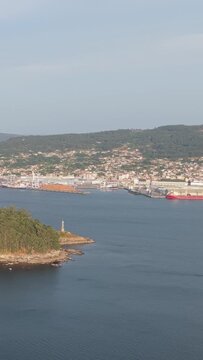 Vertical 9:16 aerial video showing the forested Illa de Tambo island in the foreground and the coastal cityscape of Combarro, Spain in the background.