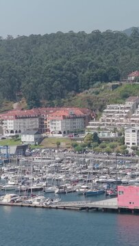 Vertical 9:16 video showing a marina filled with numerous sailboats and yachts, with residential buildings. Filmed in Combarro, Galicia, Spain.
