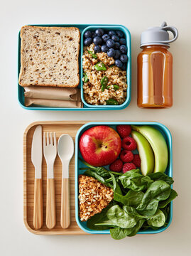 School lunch box flat lay packed lunch reusable container sandwich fruit utensil spinach bottle grain meal prepared for student break on beige surface
