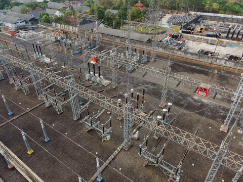 Aerial view of an electrical substation featuring steel lattice towers, power lines, and insulators set against a gravel ground in an urban area, Purwokerto Selatan, Central Java, Indonesia.