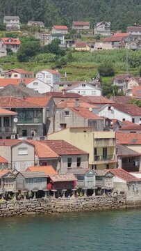 Vertical 9:16 video showing the waterfront of Combarro, Spain, with a row of traditional Galician granaries (horreos) on stone stilts along the edge of the water. The scene is bright and clear.
