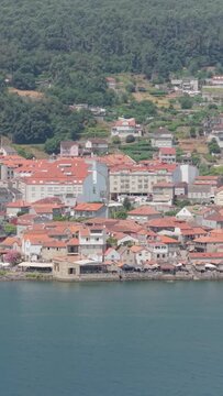 Vertical 9:16 aerial video showing the waterfront village of Combarro, Spain. The calm blue water in the foreground contrasts with the dense forest and terraced landscape behind the town.