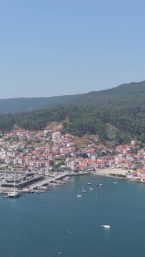 Vertical 9:16 aerial video showing the coastal village of Combarro, Spain. The calm blue waters of the Ria de Pontevedra and the picturesque Galician landscape are visible throughout the sequence.