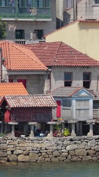 Vertical 9:16 video showing a close-up view of traditional stone granaries (horreos) and rustic houses with terracotta tiled roofs along the waterfront in Combarro, Spain.
