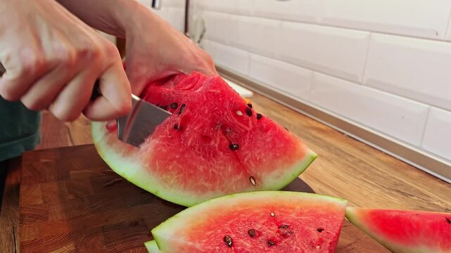 A person slices a ripe watermelon on a wooden cutting board. The watermelon has a vibrant red interior with black seeds and a green rind.