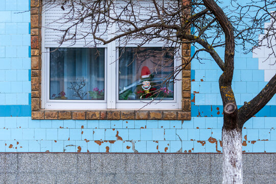 View of a Santa Claus decoration displayed in a window on a blue tiled wall with a bare tree in the foreground in Beska, Vojvodina, Serbia.