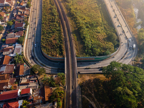 Aerial view of a railway bridge crossing a road in an urban area with houses, lush greenery, and traffic in Purwokerto, Central Java, Indonesia.