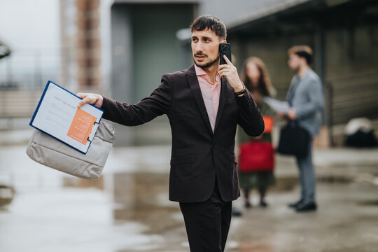A businessman in a dark suit talks on his smartphone while holding a briefcase and clipboard. He gestures toward something as two blurred coworkers stand in the background.