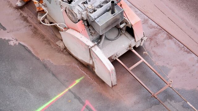 Industrial road saw cuts asphalt pavement with water cooling during construction work. Heavy cutting machine creates precise trench line while worker operates equipment on wet urban surface