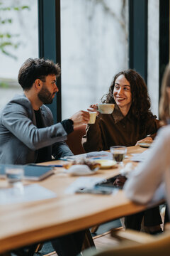 Two business colleagues share coffee and chat while seated at a meeting table. They smile and toast cups in an informal office cafe meeting.