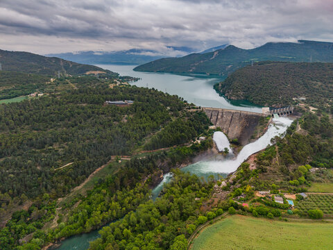 Aerial view of Panta de Sant Antoni dam on the Noguera Pallaresa River with water discharging into the valley surrounded by lush forests and Pyrenees mountains in Lleida, Catalunya, Spain.