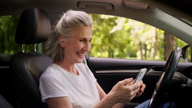 Happy excited Caucasian old woman winning with mobile phone inside automobile elderly senior female win with smartphone achieve online victory celebrate retired lady in auto successful renting car