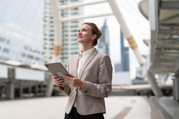 Fototapeta premium Confident businesswoman holding digital tablet looking up in the city with modern skybridge architecture, vision, technology, urban business and professional lifestyle concept.