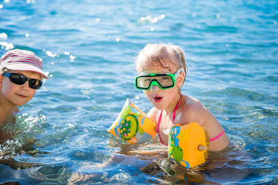 Young blond boy in striped cap and blonde girl with orange floaties swim together in clear blue sea.