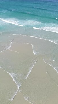 Vertical 9:16 aerial video showing turquoise waves gently washing over the sandy shoreline at Praia do Rostro, Spain.