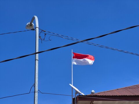Indonesian red and white national flag waving against a clear blue sky with street light, utility wires and rooftop in a tropical residential area.