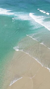 Vertical 9:16 aerial video of Praia do Rostro beach in Spain, showing turquoise waves gently washing over golden sand. The clear Atlantic water creates beautiful patterns as it meets the shoreline.