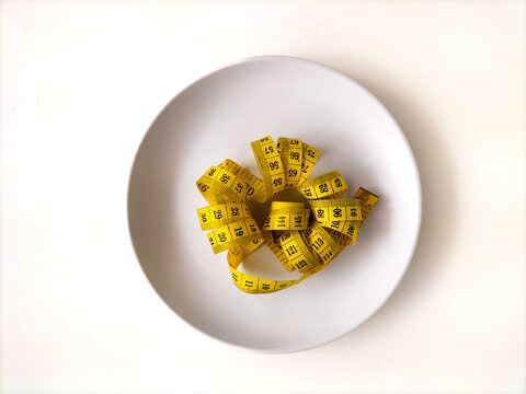 Coiled yellow measuring tape served inside a white plastic plate. This minimalistic composition visually highlights strict dieting, obesity prevention, healthy portion limits, and fitness goals