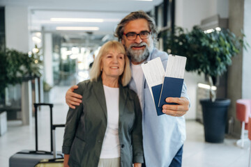 Fototapeta premium Senior couple smiling and holding travel tickets in a modern airport terminal with luggage and greenery in the background, ready for their journey