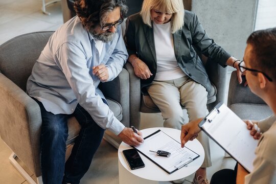 Middle-aged couple discussing paperwork with financial advisor in modern office setting, seated on gray armchairs, clipboard and smartphone on table
