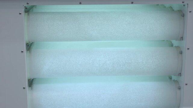 Industrial air filtration unit with three cylindrical filter elements inside pale metal housing, lit by cool cyan light, showing textured surfaces, bolts, brackets, and symmetrical technical structure