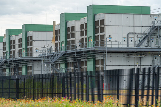 View of the industrial data center facility featuring modular cooling units, metal staircases, and a security fence with wildflowers in the foreground in Middenmeer, North Holland, Netherlands.