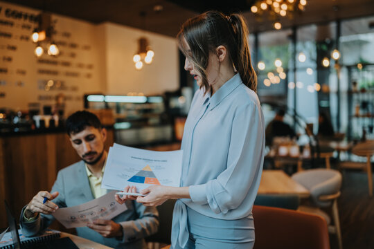 Two business colleagues review printed charts while working at a cafe table. A woman presents a chart to a seated coworker during a casual business meeting.