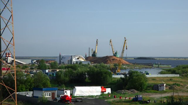 Riverside industrial port with tall gantry cranes above bulk material piles beside calm river under clear blue sky, showing storage area, shoreline, metal structures, and wide working landscape