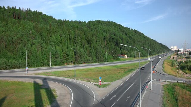 Curved elevated highway interchange beside forested hillside with vehicles traveling through merging lanes under bright midday sunlight and long lamppost shadows
