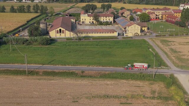 Aerial drone view of a green tractor towing a trailer across agricultural land in Lombardy, Italy, traveling toward the corn harvest during the farming season.
