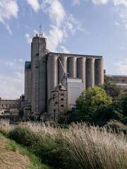 Factory building and silo complex in an industrial landscape, weathered concrete towers, metal silos, and surrounding greenery under a bright blue sky with scattered clouds © Alexander