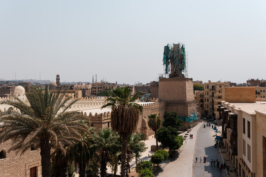 View of the historic Al-Hakim Mosque with its minaret under restoration, surrounded by palm trees and traditional architecture in the streets of El-Gamaleya, Cairo Governorate, Egypt.