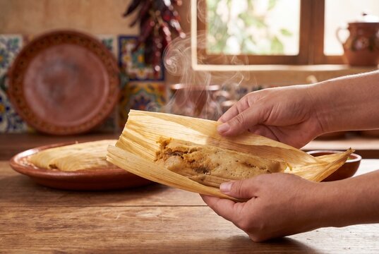 A person's hands carefully unwrapping a hot, steaming traditional Mexican tamale from its corn husk, revealing the savory filling inside, set in a rustic kitchen.
