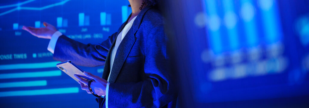 Businesswoman giving a data presentation with a tablet and digital charts on large screens, representing financial analysis, market performance, business strategy, leadership, and analytics.