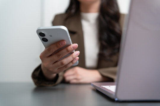 Businesswoman using a smartphone beside a laptop at an office desk, representing mobile communication, online messaging, digital workflow, business connectivity, multitasking, and modern technology.