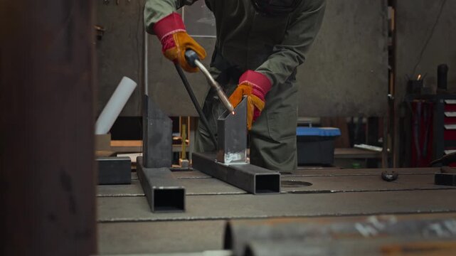 Professional welder in protective gear welding steel profiles on workbench in workshop, bright sparks flying. Clip is ideal for manufacturing, vocational training, and industrial services marketing
