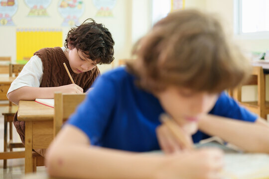 Focused elementary student writes during a classroom exam while classmates work quietly, reflecting concentration, academic assessment, and everyday school learning.