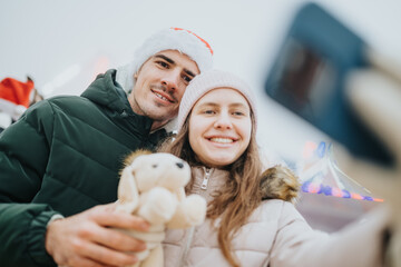 Fototapeta premium Young couple smiling for a Christmas selfie while wearing winter hats and coats. A joyful holiday selfie scene with a plush toy and festive hat.