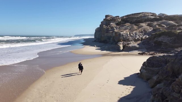 A static aerial shot capturing a couple standing together on the sandy shore, viewed from behind, looking out at the endless expanse of the Atlantic Ocean at Praia dos Coxos, Portugal.