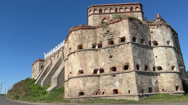 Large round fortress bastion with brick crown and stone loopholes stands next to white decorative rampart, tree and road nearby.
