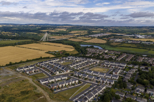 Aerial view of a modern residential neighborhood with rows of houses and dark roofs, surrounded by green fields, with the Boyne Valley Bridge in Drogheda, County Louth, Ireland.