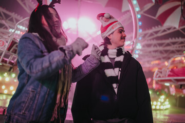 Fototapeta premium Two friends wearing winter clothes and holiday accessories enjoying a lit amusement ride. A festive nighttime carnival scene with laughter and seasonal decorations.