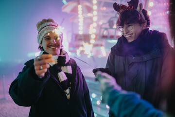Fototapeta premium Two friends hold lit sparklers and smile at an outdoor Christmas fair at night. The festive scene shows them enjoying winter lights and holiday cheer together.