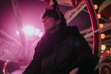 Fototapeta premium A young man wearing glasses and a winter coat smiles while riding a ferris wheel at night. He enjoys colorful carnival lights and a festive atmosphere with friends nearby.