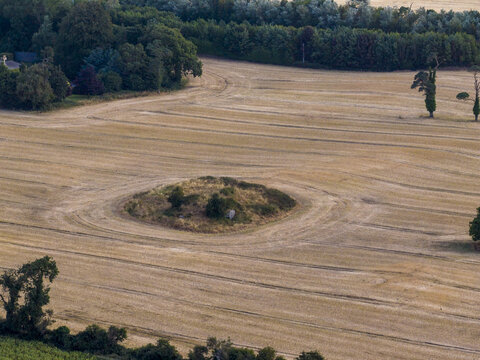 Aerial view of a historic circular tumulus mound situated in a harvested golden field with surrounding trees and rural landscape in Drogheda, County Louth, Ireland.